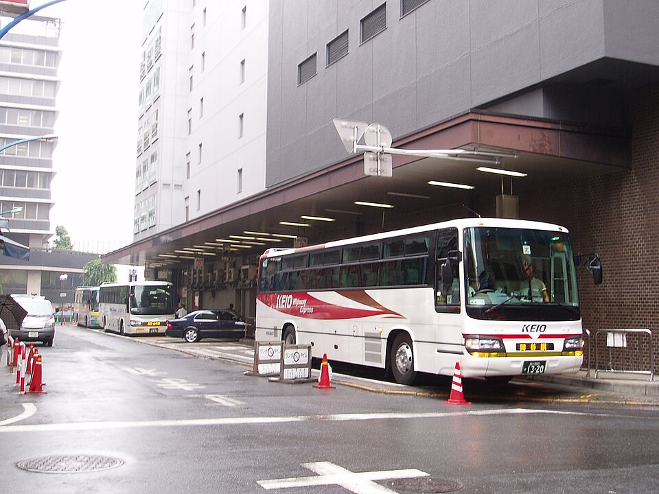 A white Keio Highway Express coach parked at a covered bus bay beside a city building, with traffic cones in the road and more coaches behind
